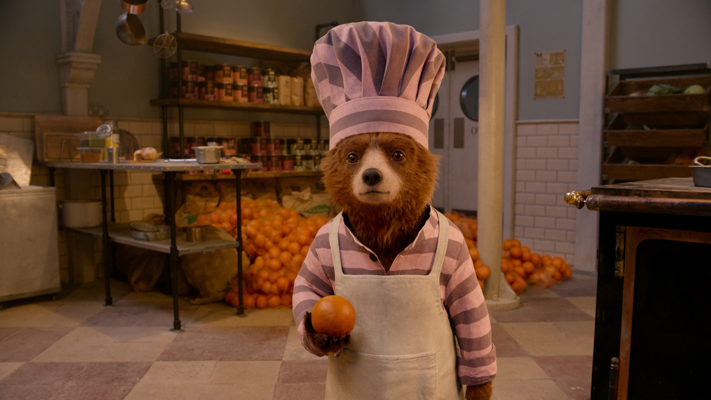 Paddington stands in a pink prisoners uniform wearing a chef's hat and holding an orange.