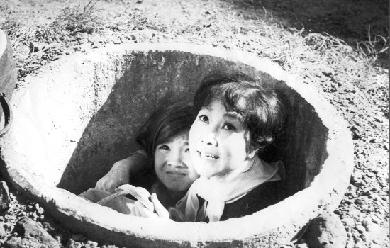 A woman and young girl crouch in a hole in the ground, looking fearful.