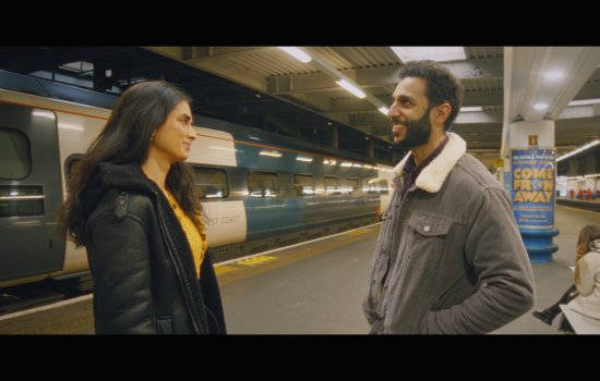 A young man and woman stand and smile at each other in the middle of a tube station.