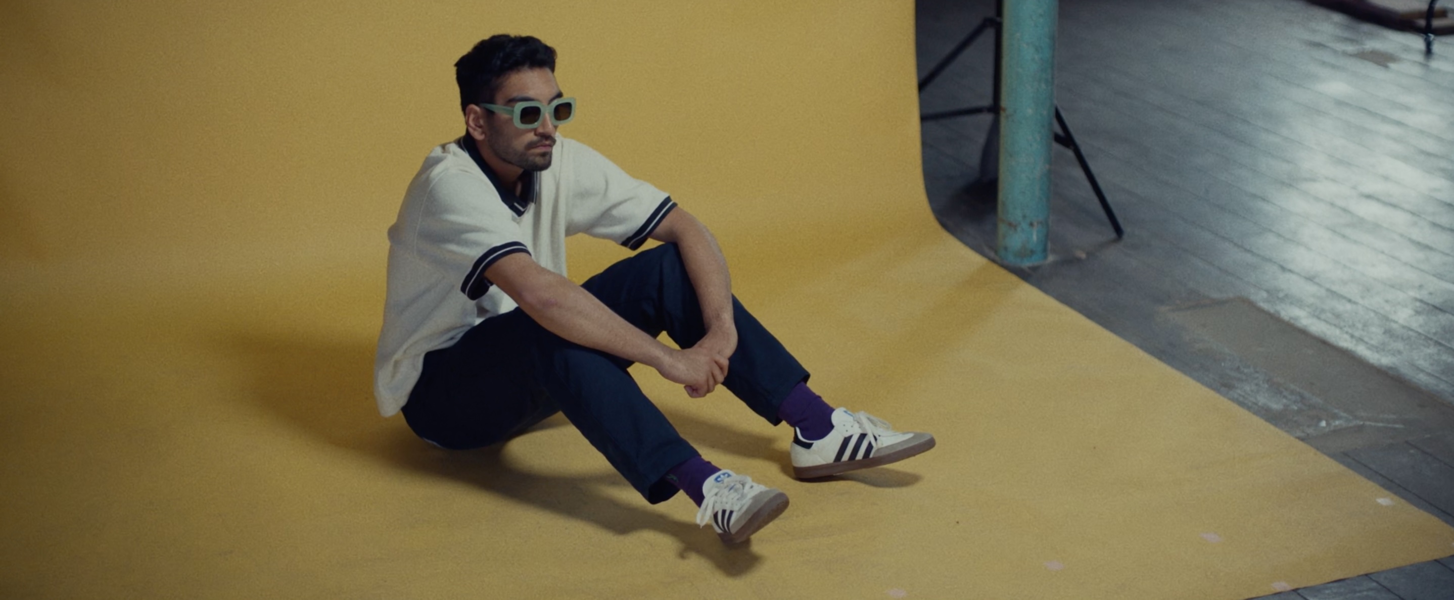 A young man wearing sunglasses sits on a yellow background.