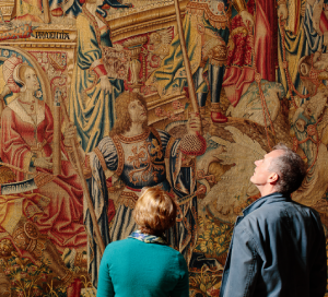 A woman wearing a green jumper and a man wearing a grey jacket stand admiring a medieval tapestry.
