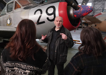 A man stands in front of an aeroplane providing a talk to two visitors listening. 