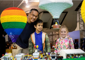 A man and two children look at a hot balloon made from LEGOs. 