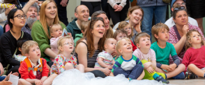 A crowd of small children and their parents are smiling while watching a performance in the grand gallery of the National Museum of Scotland. 