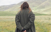 A person with long brown hair wearing a grey woolen coat walks away from the camera in the Scottish countryside