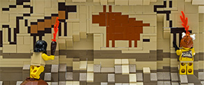 Two LEGO minifigures representing cavemen hold LEGO flame torches and look at LEGO cave paintings on a wall