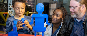 A child on the left builds a LEGO model made of blue bricks, two adults look on the right.