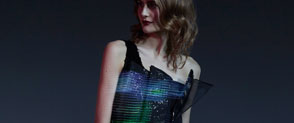 A white model looks away from the camera wearing a one-shoulder dress. On the dress are blue and green lights which are trailing across the image.