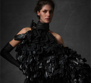 A white model looks directy at the camera with one hand on her hip, wearing a black sleeveless dress made of scraps of bin bags
