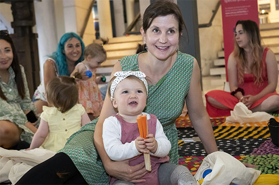 A parent with long brown hair tied back sits on a multi-coloured rug with their young baby. Both are smiling.
