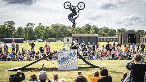 A stunt rider on a BMX bike is upside down in mid-air above a ramp outside at the National Museum of Flight. A crowd of onlookers sit on the grass taking pictures.