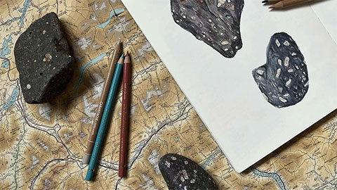 Three pencils and two pieces of rock sit on a table covered in a map. Beside them is a piece of white paper featuring pencil drawings of the rocks.