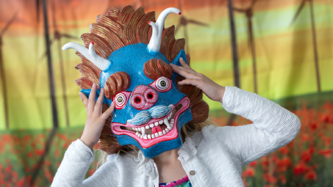 A young child wearing a white cardigan holds a a blue and pink dragon mask to their face.