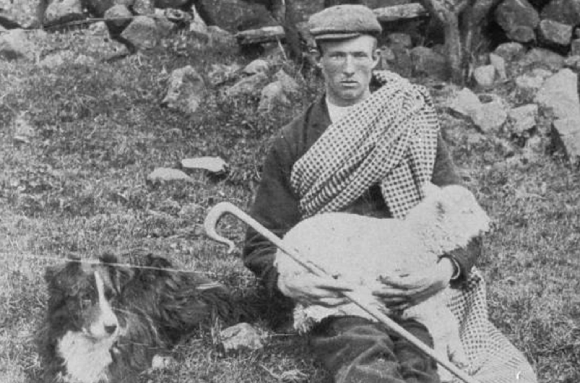 A young shepherd sits on the ground near a stone wall. He is holding a lamb on his knees. A sheepdog sits near him.