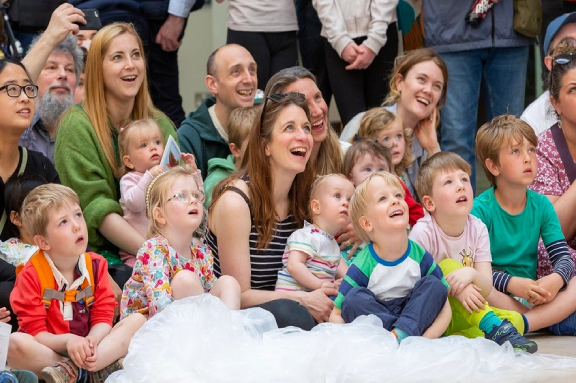 A group of children sit with their carers and parents on the floor at the museum. They're gazing up at something being performed behind the camera..
