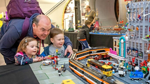 A parent and two young children standing at a table displaying a model Lego city and train models.