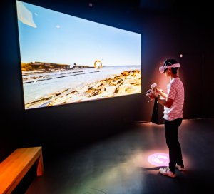 A young man stands in front of a large screen playing with a virtual reality headset