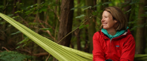 A woman wearing a red jacket sits in the woods smiling. 