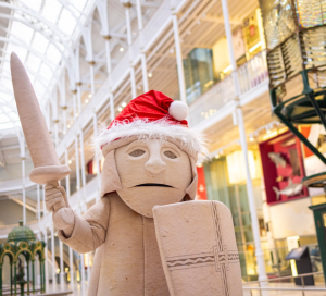 A mascot of a Lewis chess piece stands in the Grand Gallery wearing a red Santa hat, waving at the camera. 