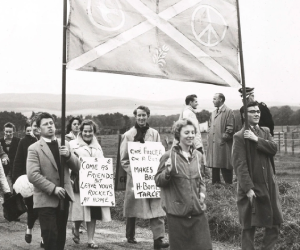 A black and white image of protesters marching underneath a peace banner. 