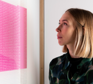 A person is looking at a framed picture. The picture looks like a square block of pink from a distance, but on closer inspection it's a stream of code for a virus.