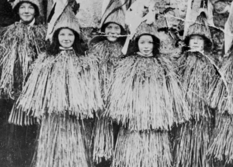 A black and white photograph of women dressed in tall coned decorative hats and flowing, straw like tops and skirts.