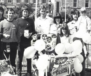  black and white photo of a group of people standing in a city square holding balloons. There is a child's pram covered in protest signs and buttons.