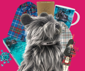 A selection of Scottish inspired gifts sit against a pink background, the items include tartan scarves and stuffed toy dog. 