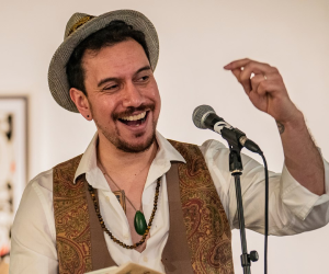 James Nokise stands in trilby hat reading from book.