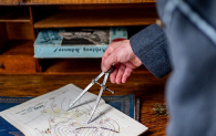 Someone is using a compass on a 1940s map. The map is placed on a desk, and the hand using the compass is visible as the person stands just out of shot, though they are wearing a blue RAF uniform.