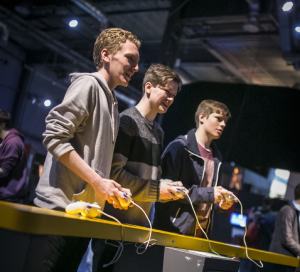 Three young men stand in front of a gaming console playing against one another. 