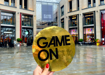 A hand holding a large golden coin with the words 'Game On' written on it, in front of the St James Quarter.