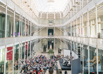 The Grand Gallery is full of natural daylight with a crowd seated, watching an orchestra playing. 