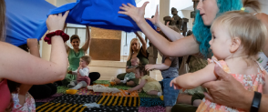 A group of women and tolders throw up a blue blanket while seated in a circle, as part of our Magic Carpet sessions.