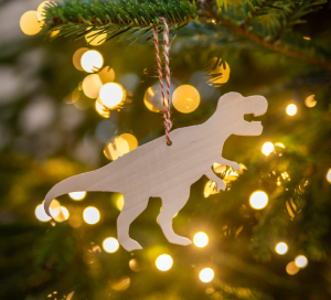 A wooden cut out of a T-Rex hangs on a Christmas Tree, it's a close up image with yellow fairy lights in the background.