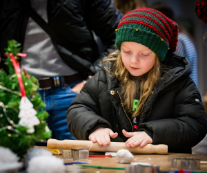 A young girl wearing a red and green Christmas hat rolls clay on wooden board. 