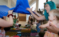 A number of young children sit with adults on a colourful carpet.