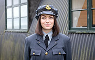 A member of museum staff with shoulder length brown hair wears replica Second World War clothing.