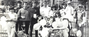 A black and white photo of a group of people standing in a city square holding balloons. There is a child's pram covered in protest signs and buttons.