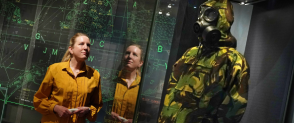 A woman wearing a yellow shirt and holding white gloves looks at a camouflage suit in Cold War Scotland