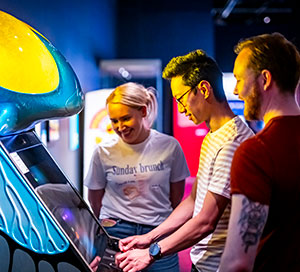 Three adults gathered around a Space Invaders arcade machine in the Game On exhibition at the National Museum of Scotland.