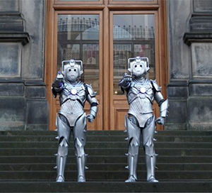 Two Cybermen stand facing you on the front steps of the National Museum of Scotland