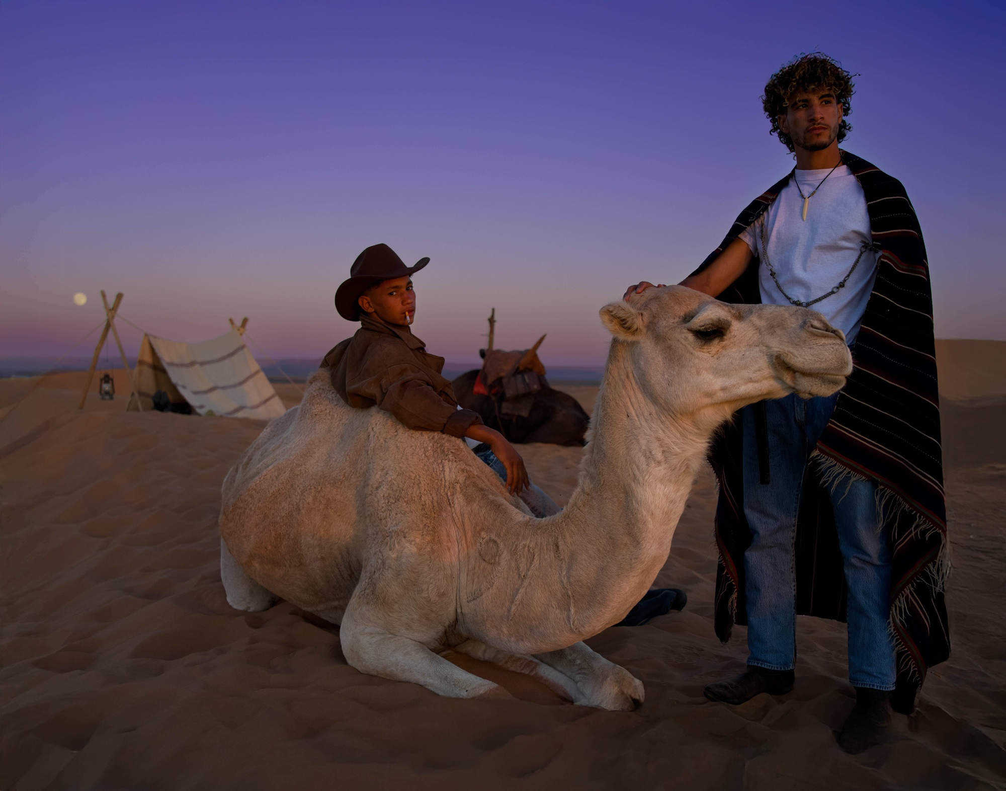 Ein Doppelporträt von zwei jungen marokkanischen Männern und einem Kamel, die im Sand gegen einen Sonnenuntergang liegen und in die Ferne schauen, eins lehnt sich gegen das Kamel mit einem Cowboyhut und rauchen einer Zigarette, der andere steht mit der rechten Hand auf dem Kamelkopf und trägt eine gemusterte Decke, den Mond und ein Zelt hinter sich. 