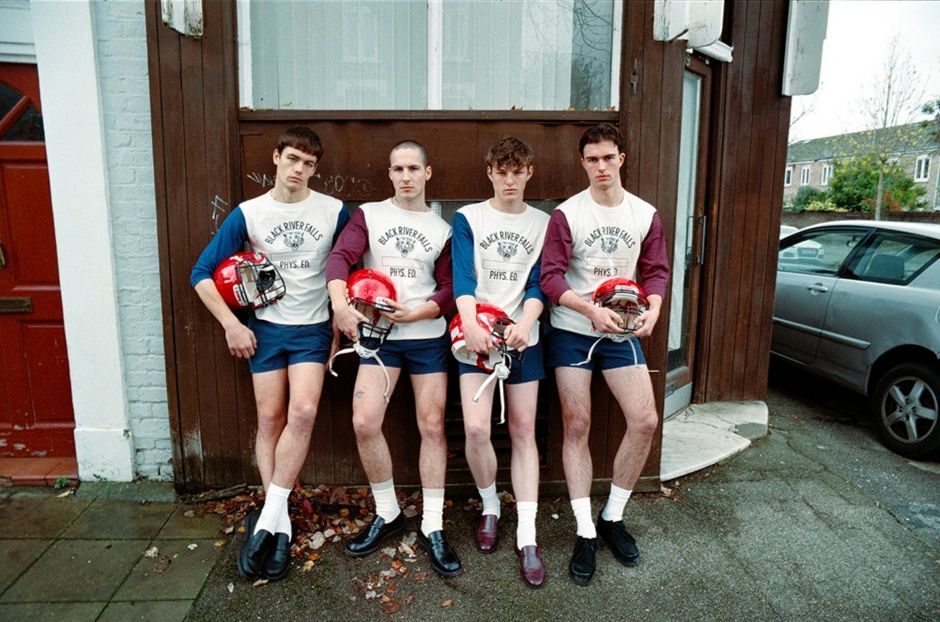 Ein Gruppenporträt von vier weißen jungen Männern, die vor einem holzgetäfelten Eckhaus sitzen, stehen sie der Kamera gegenüber und tragen American-Football-Helments, die weiße T-Shirts und kurze blaue Shorts, weiße Knöchelsocken und Loafer aus Lackleder tragen. 