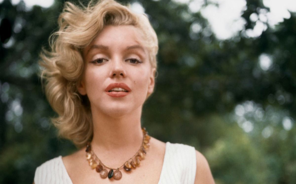 Ein Farbfoto einer weißen Frau von der Taille aufwärts. Sie hat kurzes blondes Haar, das vom Wind zur Seite weht. Sie hat leicht geöffnete rote Lippen und trägt eine weiße gerippte Weste sowie eine braune Perlenkette. Sie steht draußen, mit verschwommenem Grün hinter sich.
