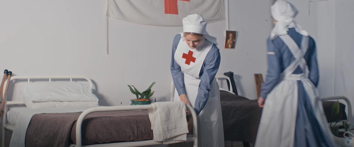 Women wearing replica WWI Nurse uniforms, making a bed in a set made to look like a WWI-era hospital