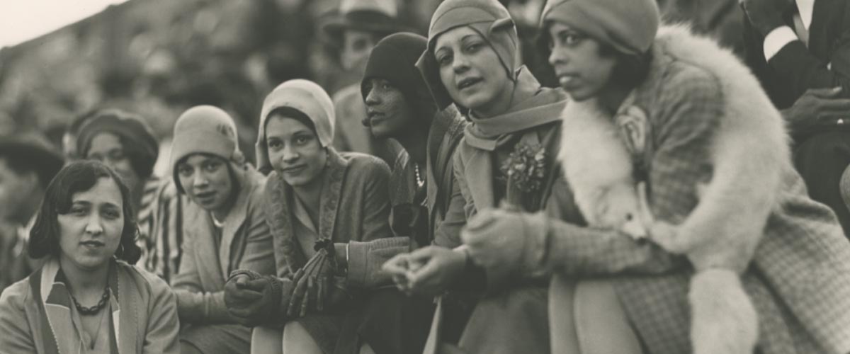 A group of African American flappers: young women dressed in 1920s clothing including coats, gloves, close-fitting cloche hats and one in the foreground with a full light colored mink or fox stole. They are sitting on what appears to be bleachers in front of a larger crowd.