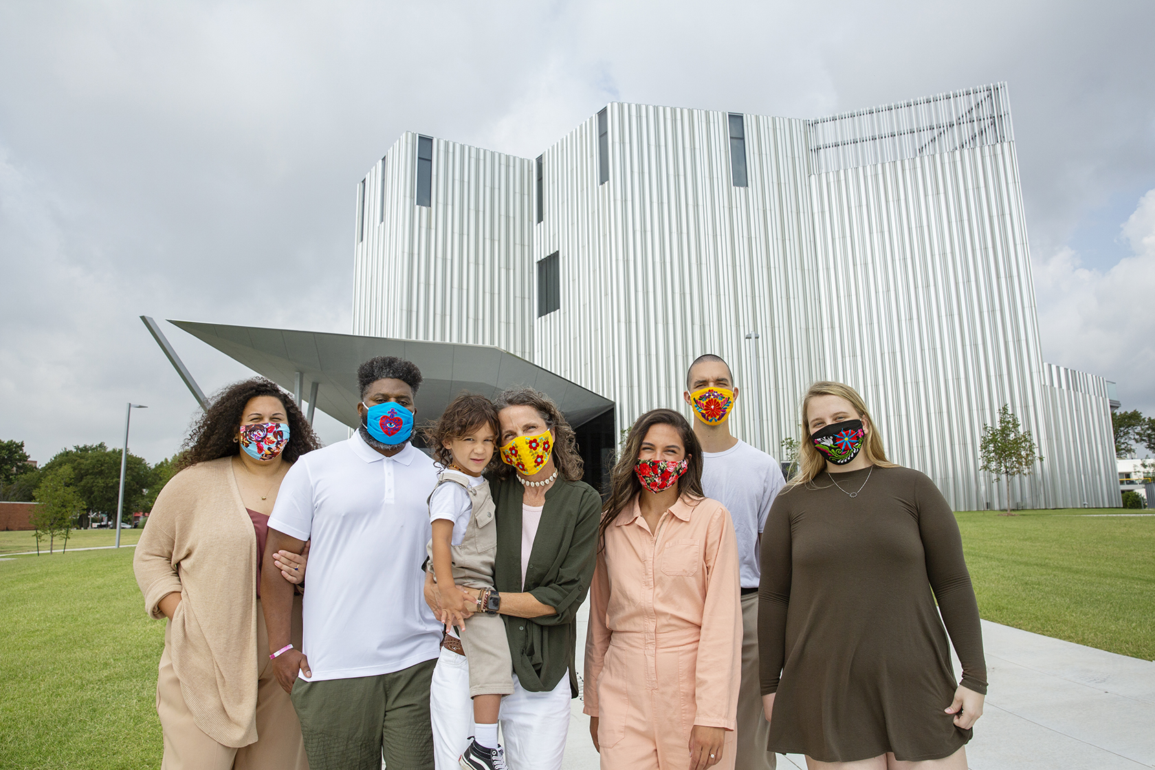 Several people wearing brightly colored face coverings stand together in front a tall silver aluminum building.
