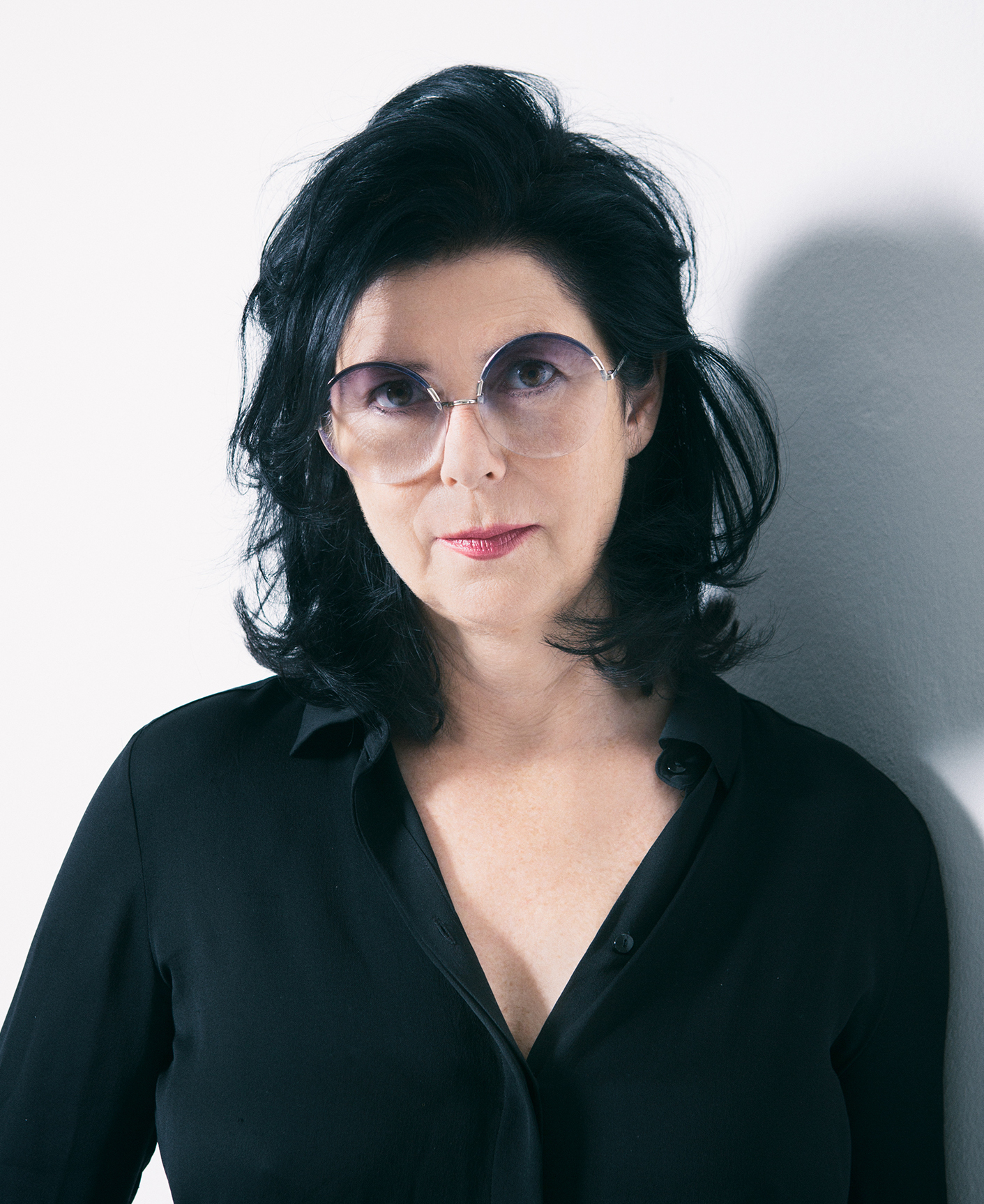 A woman in a black top with black hair wearing sunglasses in front of a white wall.