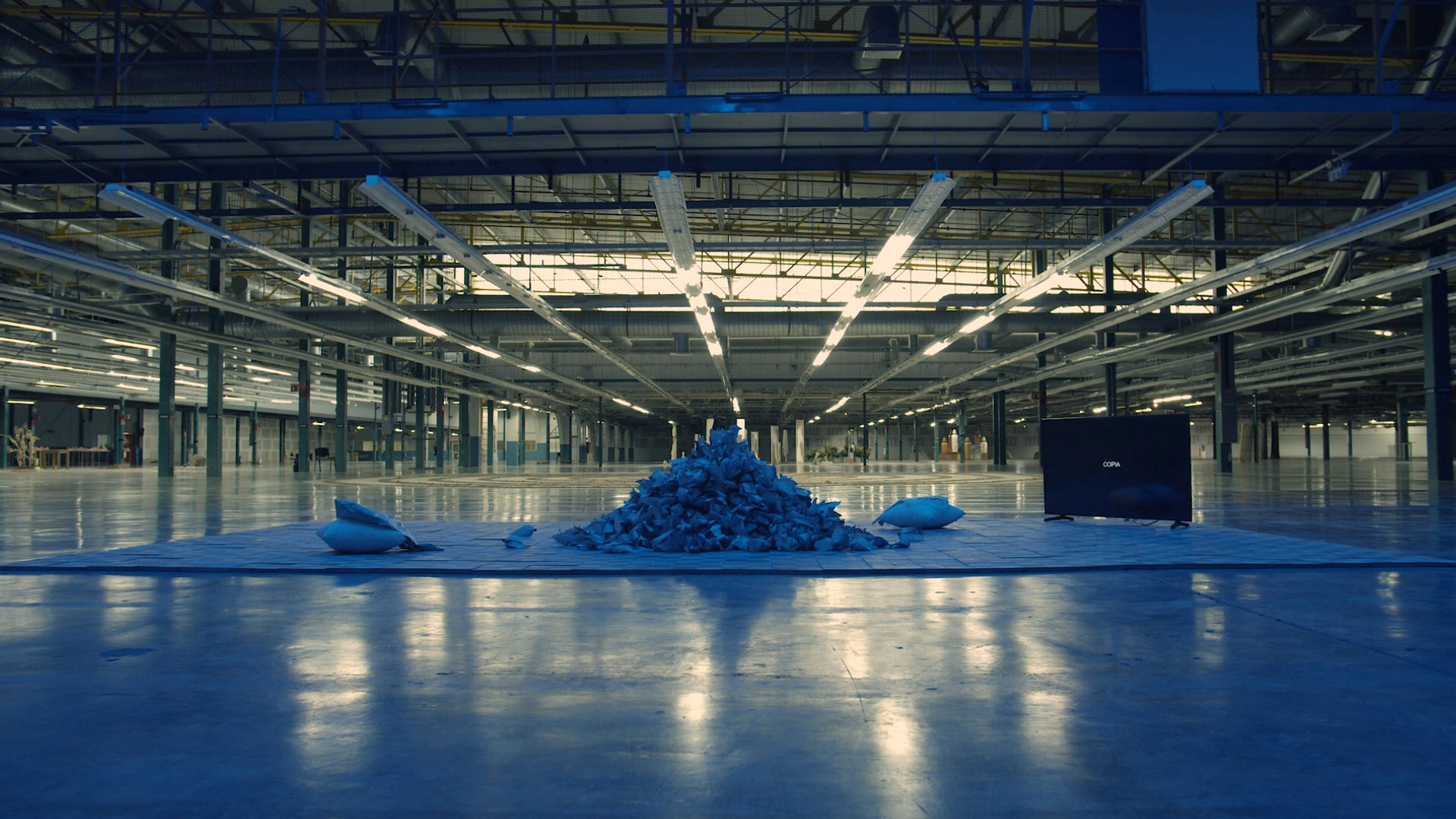 An art installation featuring a mountain of folded and crumpled paper next to a large television screen in an empty, blue-lit warehouse space